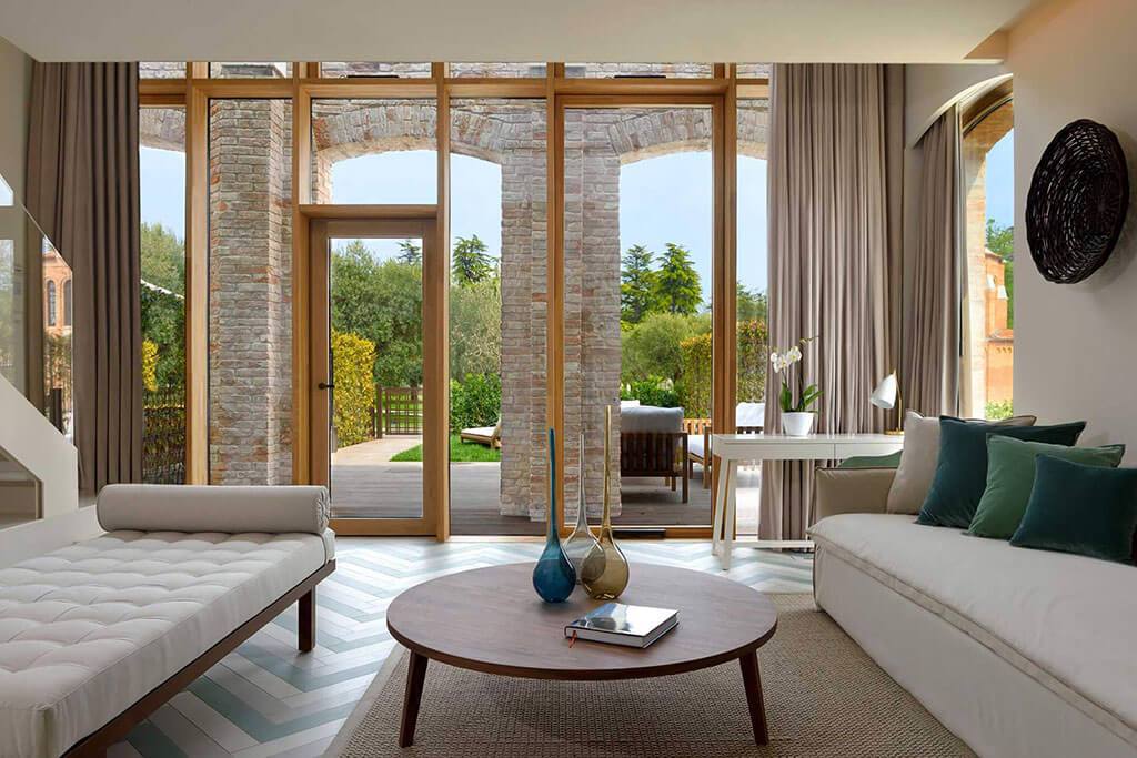 Okna drewniano – aluminiowe, kolor naturalny dąb. Widok z wewnątrz obszernego apartamentu na wyjście na taras i ogród. Ściana w postaci przeszklenia z przeszklonymi drzwiami i dużym oknem przesuwnym.