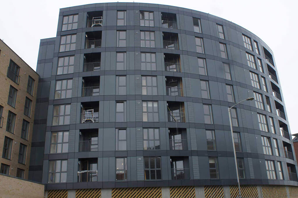 Okna drewniano – aluminiowe, kolor antracyt, widok na front budynku wielopiętrowego, okna balkonowe, duże przeszklenia. Bryła budynku zaokrąglona